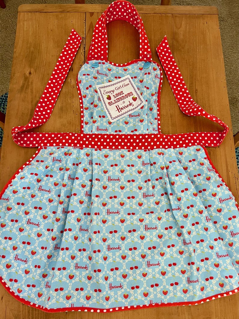 May include: A red and white polka dot apron with a blue and white patterned skirt. The skirt has a repeating pattern of strawberries and the Harrods logo. The apron has a small white tag that says "Every Girl Can Look Glamorous at Harrods".