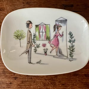 May include: A white ceramic dish with a hand-painted design of a man and a woman in front of a house. The man is holding an umbrella and the woman is wearing a pink dress. The dish has a green and white striped window and a green bush.