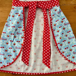May include: A blue and white apron with a red and white polka dot bow tie. The apron has a  "Harrods" logo repeated on the fabric.