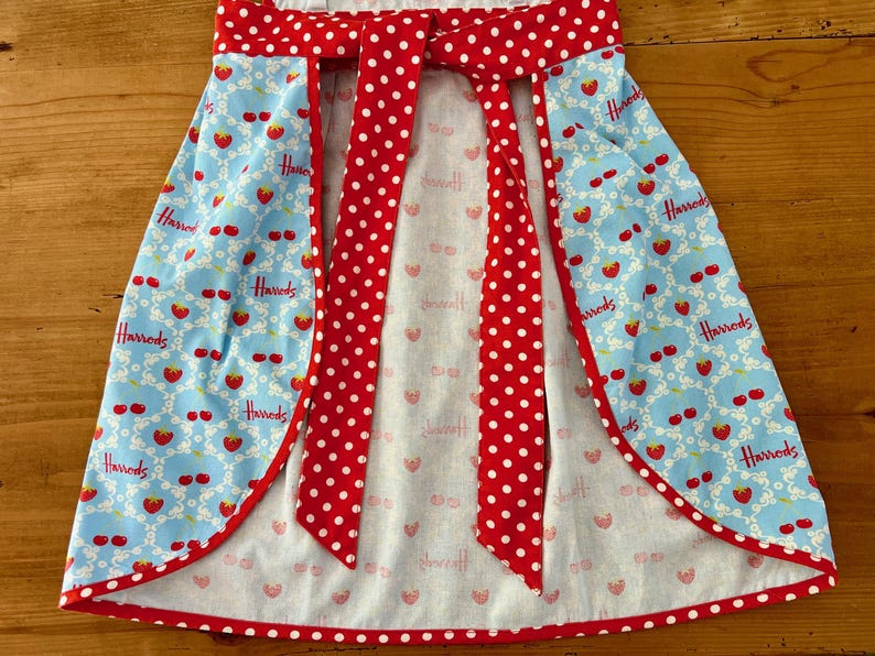 May include: A blue and white apron with a red and white polka dot bow tie. The apron has a  "Harrods" logo repeated on the fabric.