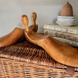 May include: Two vintage wooden shoe forms with a light brown finish rest on a woven wicker basket. The forms have a rounded heel and a long, pointed toe. A stack of books and small white plates are in the background.