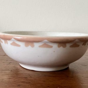 May include: A white ceramic bowl with a decorative peach-colored trim. The trim features a repeating pattern of stylized shapes. The bowl is set on a wooden surface, showcasing its simple yet elegant design.