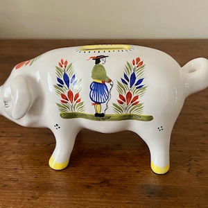 May include: A white ceramic piggy bank featuring a hand-painted illustration of a person in traditional attire standing between two floral motifs. The pig has yellow-tipped feet, a curled tail, and a coin slot on its back with yellow detailing.