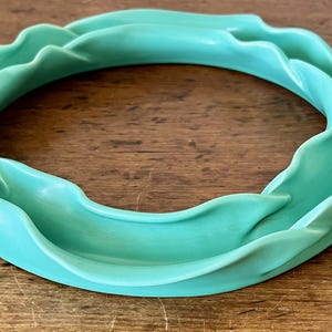 May include: A turquoise ceramic decorative ring with a wavy, organic design. The ring is circular, with a smooth, glossy finish. The object is set against a wooden surface.