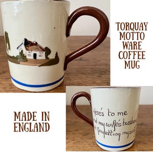 May include: White ceramic coffee mug with a brown handle. The mug has a blue stripe around the top and bottom. The front of the mug has a painted image of a small house with a red roof and green trees. The back of the mug has brown text that reads "Here's to me and my wife's husband not forgetting myself". The bottom of the mug has brown text that reads "Torquay Motto Ware Coffee Mug Made in England".