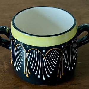 May include: A black ceramic bowl with two handles, featuring a yellow band around the rim and decorative white and gold accents. The interior is white. A decorative kitchenware item.