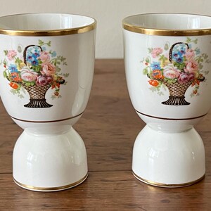 May include: Two white ceramic egg cups with a gold rim and a floral basket design. The basket contains pink, blue, and orange flowers. The egg cups have a pedestal base.