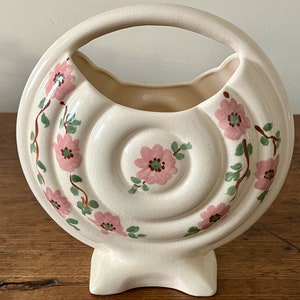 May include: Cream-colored ceramic vase with a circular design and a handle. The vase features pink floral patterns with green leaves and brown stems. The base is a rectangular shape.