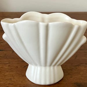 May include: A cream-colored ceramic vase featuring a scalloped, shell-like design with vertical ridges radiating from a central pedestal base. The opening has a wavy, undulating rim that follows the fluted pattern of the body.