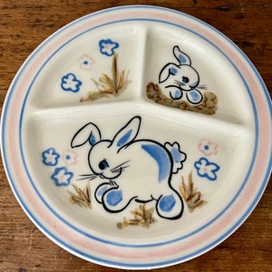 May include: A white ceramic divided plate with a pink rim. The plate features a blue and white bunny illustration with flowers and grass.