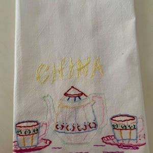 May include: A white cotton tea towel featuring hand-embroidered designs in various colors. The word CHINA is stitched in yellow thread above a central teapot flanked by two teacups on saucers, all outlined in red, blue, green, and pink thread.