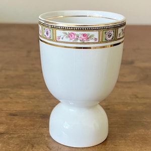 May include: White ceramic egg cup with a decorative band featuring pink roses, green accents, and gold trim. The egg cup has a rounded base and a wider top, creating a classic, elegant design. The gold trim adds a touch of sophistication.