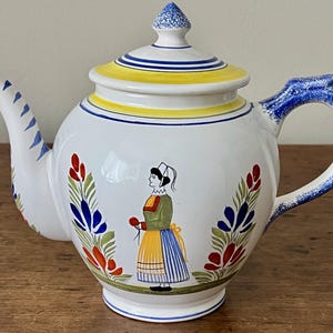 May include: White ceramic teapot with a blue handle and spout, and a yellow and blue lid. The teapot features a hand-painted design of a woman in traditional clothing, surrounded by floral motifs in red, blue, and green.