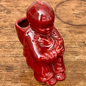 May include: A glossy, red ceramic figurine of a person carrying a basket. The figure is in a seated position, with a detailed depiction of clothing and facial features. The basket on the back has an opening, suggesting it could be used as a small planter or decorative item.