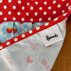 May include: A blue and white fabric with a red and white polka dot trim. The fabric has a small strawberry print. A white tag with the word "Harrods" in black lettering is attached to the fabric.