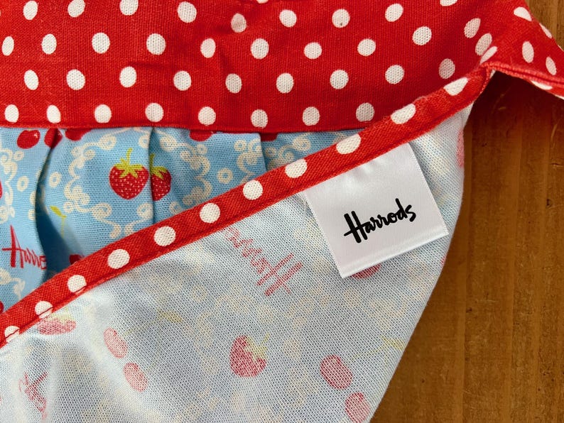 May include: A blue and white fabric with a red and white polka dot trim. The fabric has a small strawberry print. A white tag with the word "Harrods" in black lettering is attached to the fabric.