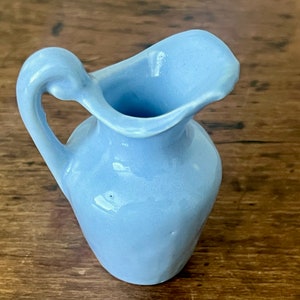 May include: A small, light blue ceramic pitcher with a handle. The pitcher has a rounded body and a flared lip.