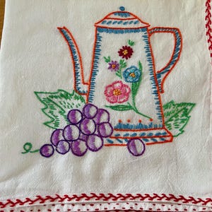 May include: White embroidered tea towel featuring a teapot design with flowers and grapes. The teapot is outlined in orange and blue, with a floral arrangement. The grapes are purple, and the towel has a red stitched border.