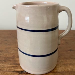 May include: A beige ceramic pitcher with a handle and spout. Two dark blue bands encircle the body of the pitcher. The pitcher is sitting on a wooden surface.