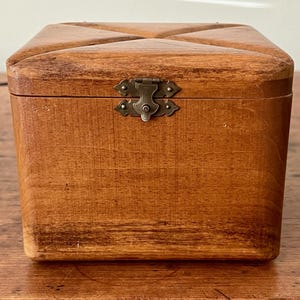 May include: A small, square wooden box with a hinged lid and a decorative metal clasp. The box is made of light brown wood and has a cross-shaped design on the lid. The box is sitting on a wooden surface.