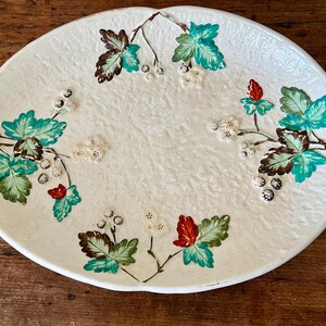 May include: Oval, off-white ceramic serving platter with embossed texture. The platter is decorated with hand-painted botanical designs featuring green, teal, and brown leaves, white berries, and red accents. The platter has two handles.