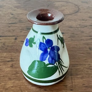 May include: A small, hand-painted ceramic vase with a brown rim and a white body. The vase is decorated with blue floral designs and green leaves. The base and top are outlined with green stripes. The vase is approximately 5 inches tall.
