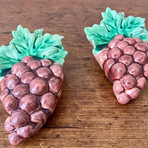 May include: Two ceramic grape clusters with green leaves. The grapes are a deep purple color, and the leaves are a bright green. The clusters are arranged on a wooden surface.