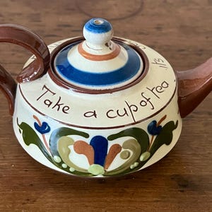 May include: A ceramic teapot with a brown handle and spout, and a lid with blue and white stripes. The teapot is cream-colored with the words "Take a cup of tea" written on it, and floral designs.