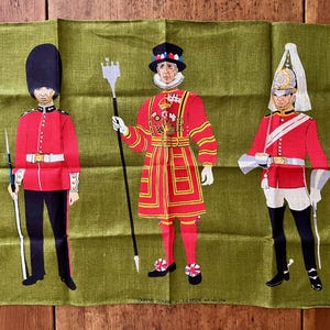 May include: A green textile featuring three illustrations of British guardsmen in red uniforms. The guardsmen are depicted with detailed attire, including tall hats and ceremonial weapons. The textile has text at the bottom.