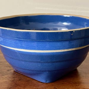 May include: A blue ceramic bowl with a cream-colored rim and a white stripe. The bowl has a square base and a rounded top. The bowl is sitting on a wooden surface. The bowl is likely used for food preparation or serving.