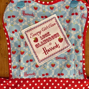 May include: A red and white polka dot apron with a blue and white floral pattern. The apron has a patch sewn on the front that says "Every Girl Can Look Glamorous At Harrods".