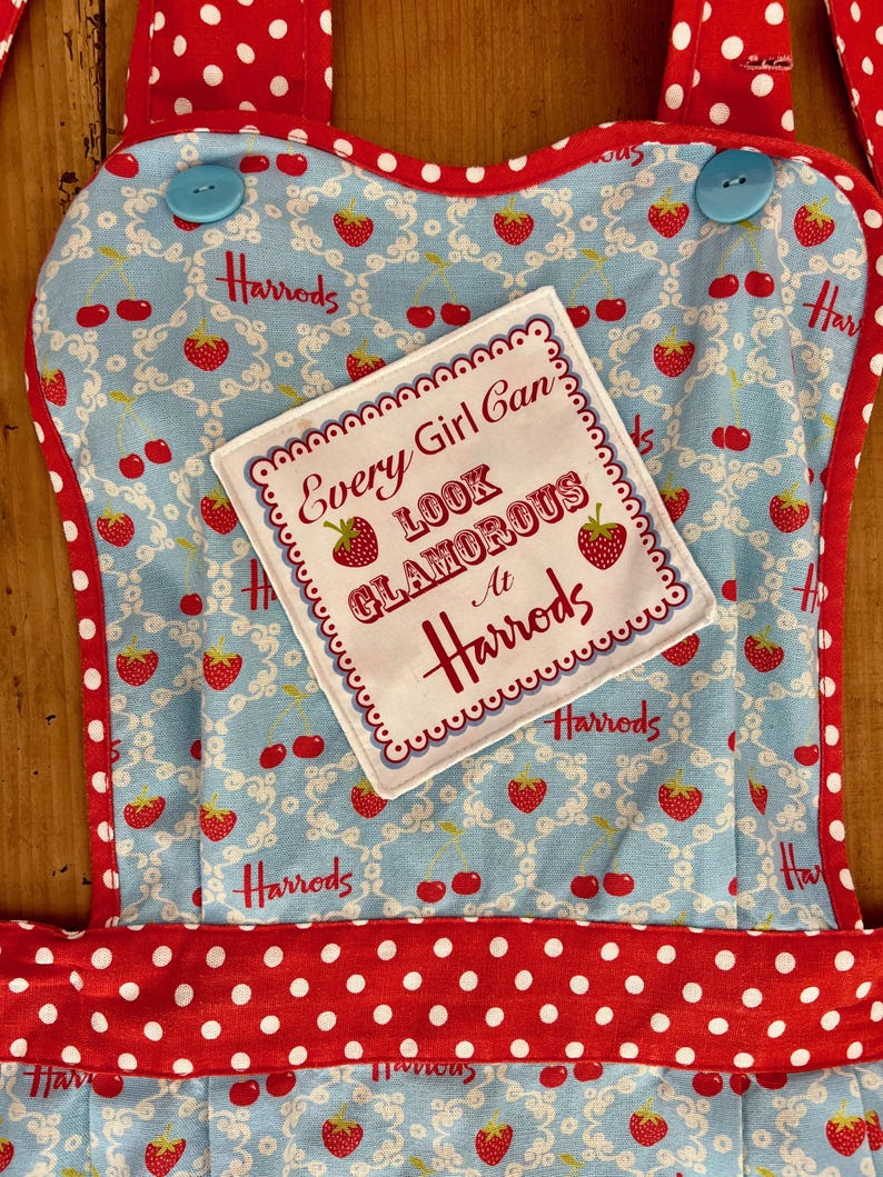 May include: A red and white polka dot apron with a blue and white floral pattern. The apron has a patch sewn on the front that says "Every Girl Can Look Glamorous At Harrods".