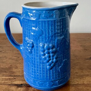 May include: A blue ceramic pitcher with a white rim and a grapevine design. The pitcher has a textured surface and a handle.