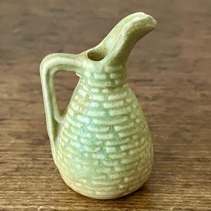 May include: A small, light green ceramic pitcher with a handle and a spout. The pitcher has a textured, brick-like pattern and is likely handmade. The overall design is simple and rustic, suitable for decorative use or serving small portions.