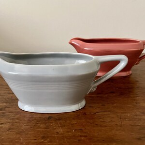 May include: Two ceramic gravy boats with a smooth, glossy finish. One boat is light gray and positioned in the foreground, while the second boat is a muted coral pink and sits slightly behind it. Both feature a wide spout and a side handle.