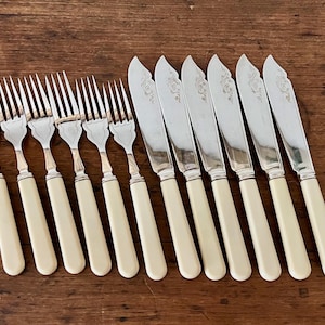 May include: A set of antique silverware, including forks and butter knives, arranged on a wooden surface. The forks have silver tines and cream-colored handles. The butter knives feature a floral design on the blades and matching handles.