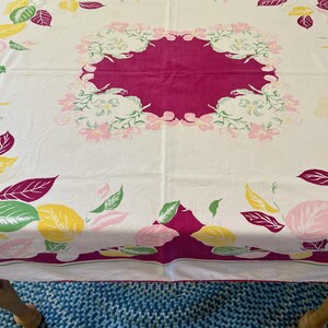 May include: A square tablecloth with a white base featuring a central magenta square surrounded by floral motifs in pink and green. The border is decorated with a pattern of scattered leaves in shades of yellow, magenta, pink, and green.