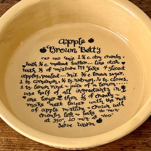 May include: A yellow ceramic pie plate with a handwritten recipe for Apple Brown Betty. The recipe includes ingredients like brown sugar, cinnamon, nutmeg, and cloves. It also instructs to bake at 350 degrees Fahrenheit for 10 minutes and then at 400 degrees Fahrenheit for 40 minutes.