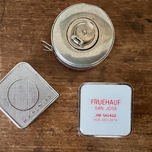 May include: An overhead shot of three vintage measuring tools. A silver metal tape measure, a silver diamond-shaped object with the text "H.K. PORTER", and a white and silver square with the text "FRUEHAUF SAN JOSE" and a phone number.
