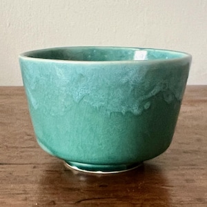 May include: A small, round ceramic bowl with a vibrant teal glaze. The surface features a mottled, textured finish with variations in color intensity, ranging from deep forest green to lighter seafoam tones, creating a unique, organic appearance.