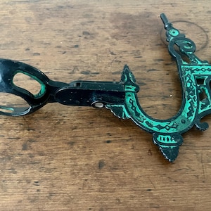 May include: An antique black and green metal ice tongs and bottle opener. The ice tongs have a circular handle and a hinged design. The bottle opener has an ornate, decorative design with green accents. The items are on a wooden surface.