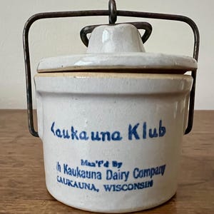 May include: A vintage stoneware crock with a lid and metal handle. The crock is off-white with blue lettering that reads "Kaukauna Klub" and "Kaukauna Dairy Company, CAUKAUNA, WISCONSIN".