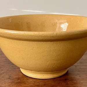 May include: A round, yellow-toned ceramic bowl with a wide rim and a slightly textured surface. The bowl sits on a small, circular base. The bowl is likely used for mixing or serving food.
