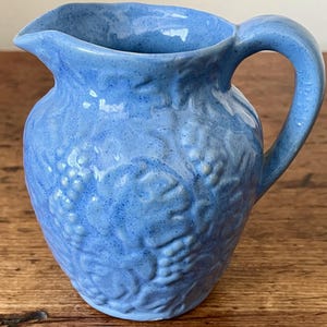 May include: A small, light blue ceramic pitcher with a textured, embossed design of grapes and leaves. The pitcher has a rounded body and a handle.