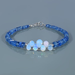 May include: A bracelet with blue gemstone beads and a central cluster of white and blue opals. The bracelet is secured with a silver clasp. The word "Vatsla" is printed on the image. The blue beads are approximately 0.3 cm in diameter.