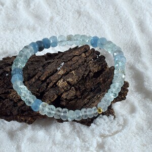 May include: A light blue and dark blue gemstone bracelet with a gold bead accent. The bracelet is made of faceted beads and is displayed on a piece of driftwood.
