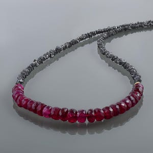 Peut inclure: Un collier composé d'une rangée de perles de rubis facettées rouge foncé au centre, reliée à une longue chaîne de petites perles de diamant noir brut. Le design met en valeur le contraste entre les pierres rouges polies et les pierres noires texturées, assemblées par de petits éléments métalliques dorés.