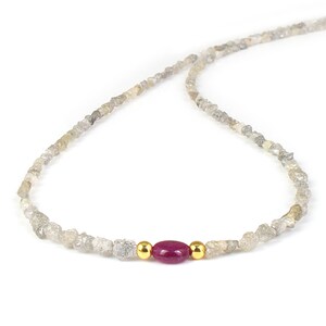 May include: A delicate necklace featuring a strand of rough-cut, gray-toned diamonds. The necklace is accented with a vibrant, oval-shaped ruby and two small gold beads. The design is simple and elegant, suitable for everyday wear.