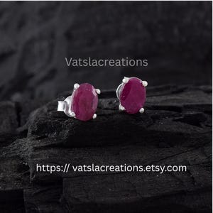 Op de afbeelding: Een paar oorknopjes met ovale, donkerroze robijnen in zilveren zettingen. De tekst Vatslacreations en de URL https://vatslacreations.etsy.com zijn zichtbaar in de afbeelding. De edelstenen hebben een glad en gepolijst oppervlak.