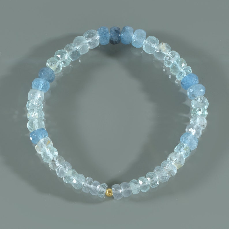 May include: A light blue aquamarine gemstone bracelet with a gold bead accent. The bracelet is made of round beads that are graduated in size, with the larger beads at the center of the bracelet.